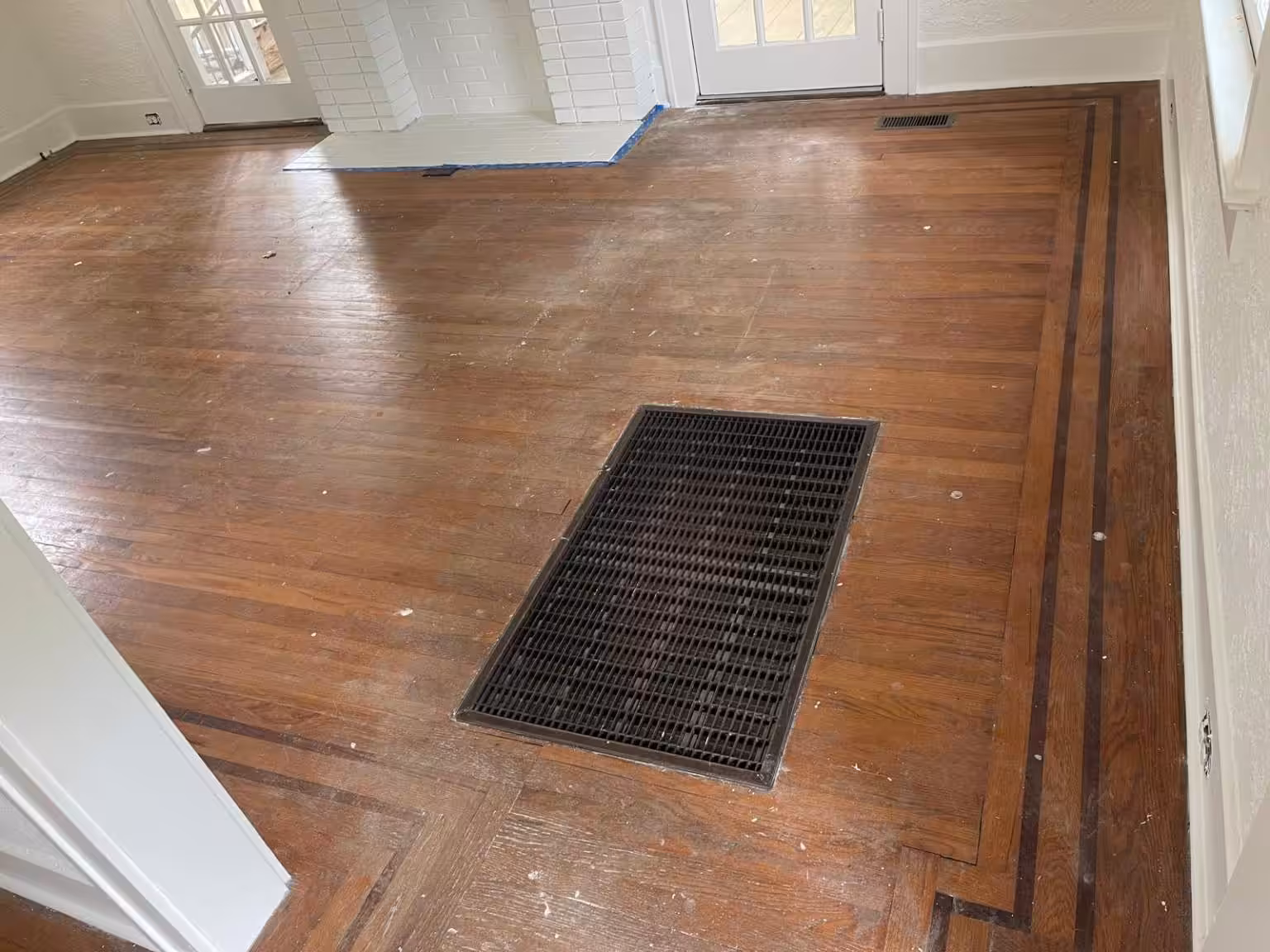 Older living room before refinishing: narrow wood strip floor with a dark border inlay, a large cast-iron floor register and a white painted brick fireplace